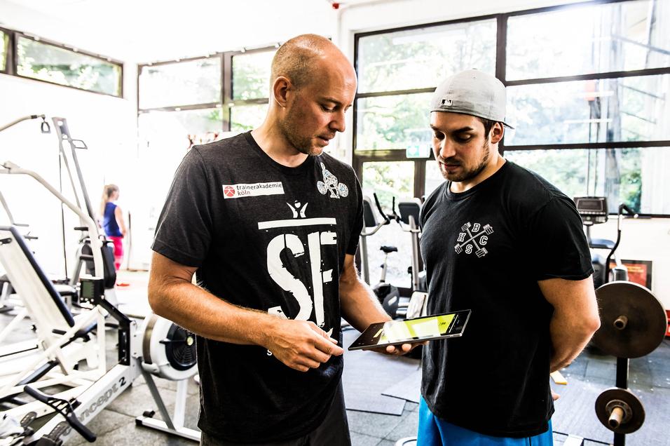 Zwei Trainer im Fitnessstudio betrachten gemeinsam ein Tablet. Im Hintergrund sind Trainingsgeräte zu sehen.